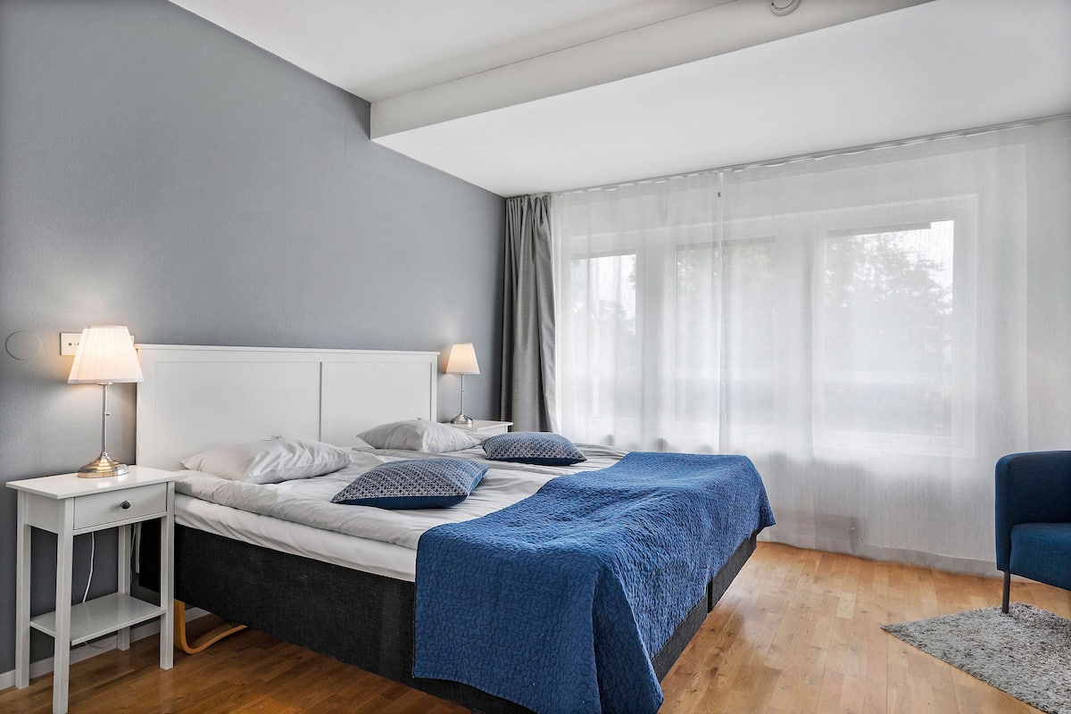 A bedroom with a bed, two bedside tables, a chair, and a window with sheer curtains.