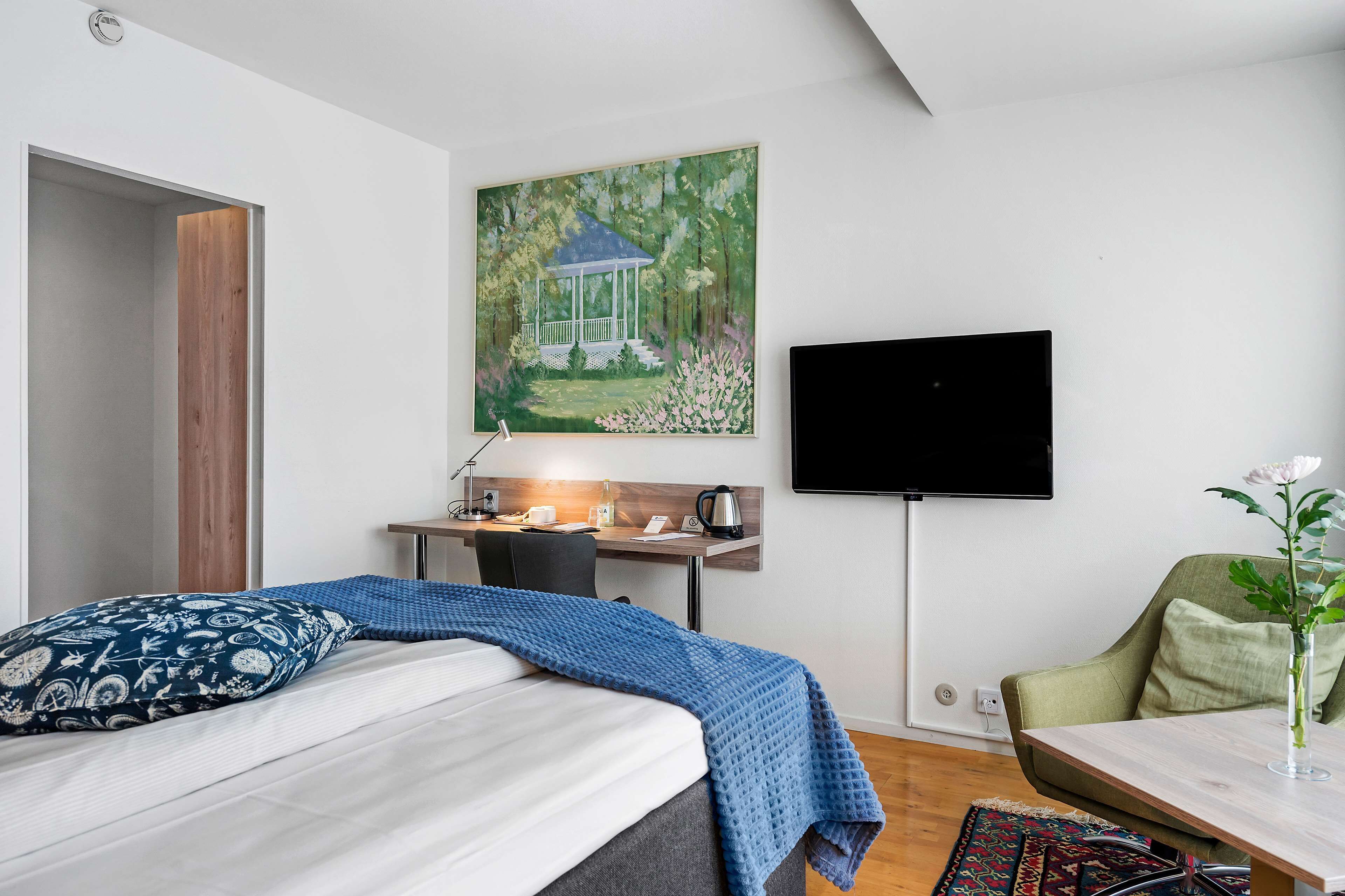 Une chambre à coucher avec un lit, un bureau avec un ordinateur, une chaise, une télévision et un tableau représentant un kiosque.