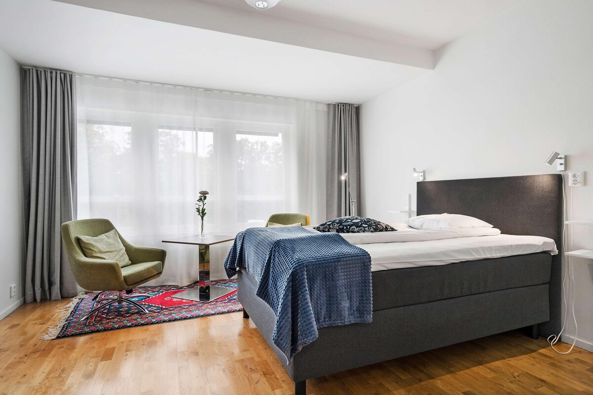 A modern bedroom with a bed, a green armchair, a small table, and a window with sheer curtains.