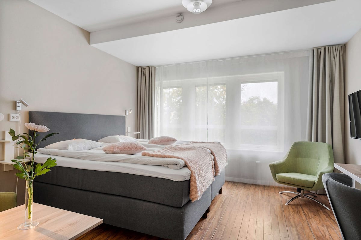 A modern hotel room with a large bed, a green armchair, a wooden table, and a vase with flowers.