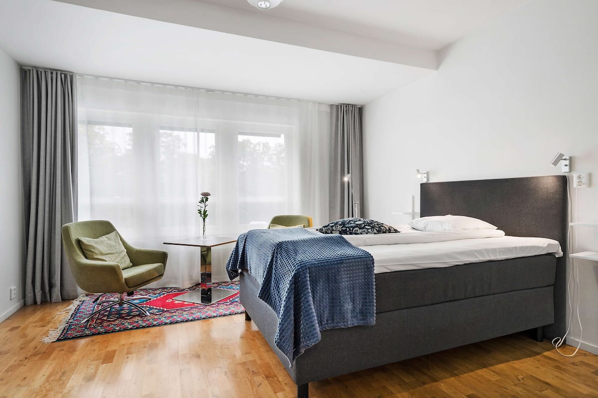 A modern bedroom with a bed, a green armchair, a small table, and a window with sheer curtains.