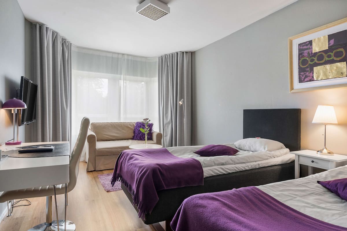 A hotel room with a desk, two beds, a TV, and a painting on the wall.