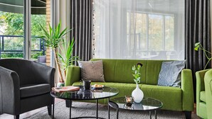 Una sala de estar moderna con un sofá verde, un sillón negro y una mesa de centro de vidrio. Hay una planta sobre el sofá y un jarrón con flores en la mesa de centro.