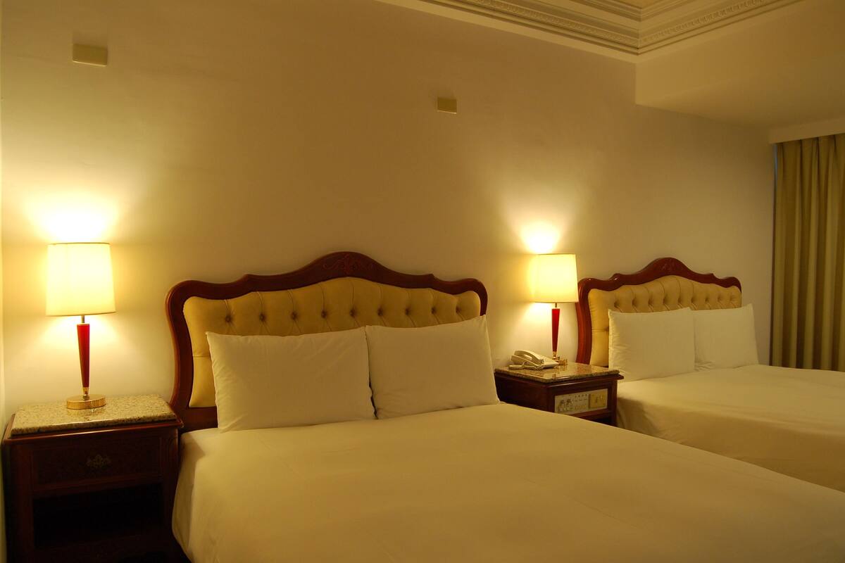 Habitación de hotel con dos camas, cada una con cabecero de madera y ropa de cama blanca. Se observan mesitas de noche con lámparas y un teléfono.