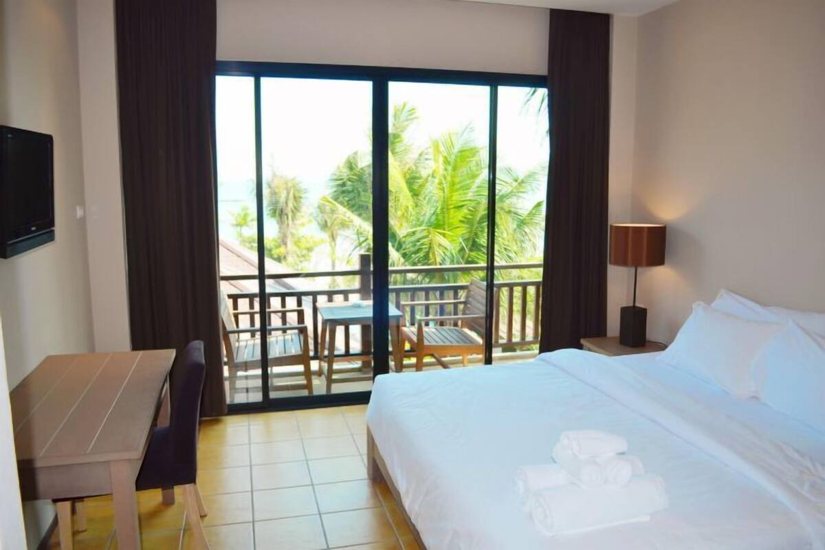 A hotel room with a bed, a desk, a chair, a table, a balcony with a view of palm trees, and a lamp.