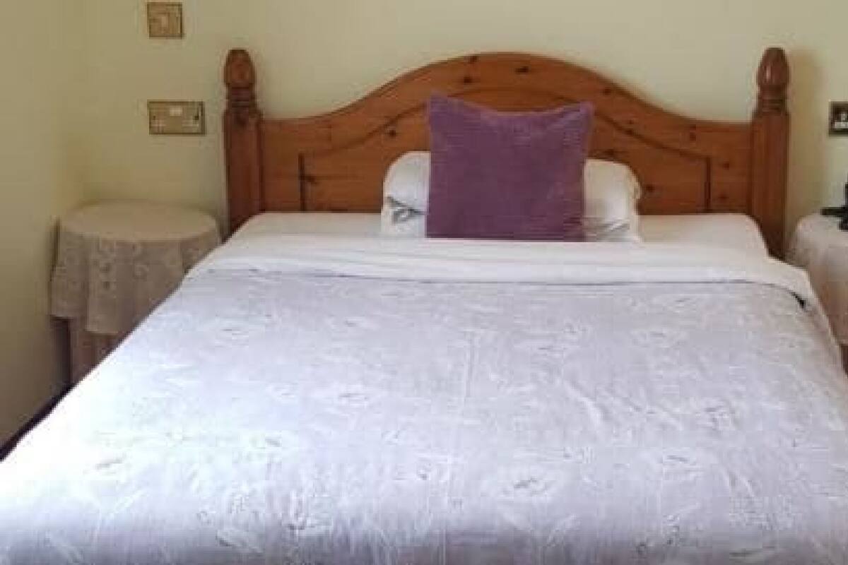 A neatly made bed with a wooden headboard, a white bedspread, and a purple pillow. There are two bedside tables with lamps and a telephone on one of them.