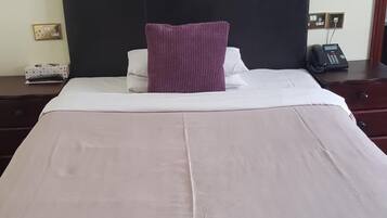 A neatly made bed with a white and pink bedspread, a single purple pillow, and a wall-mounted painting of blue flowers.