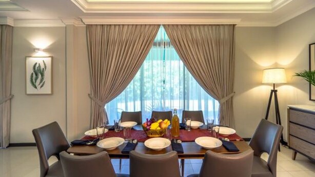 Un comedor con una mesa dispuesta para cuatro, ventanas grandes con cortinas y una lámpara a un lado.