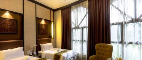 A hotel room with two beds, a ceiling fan, large windows with curtains, a small table with a fruit bowl, and artwork on the walls.