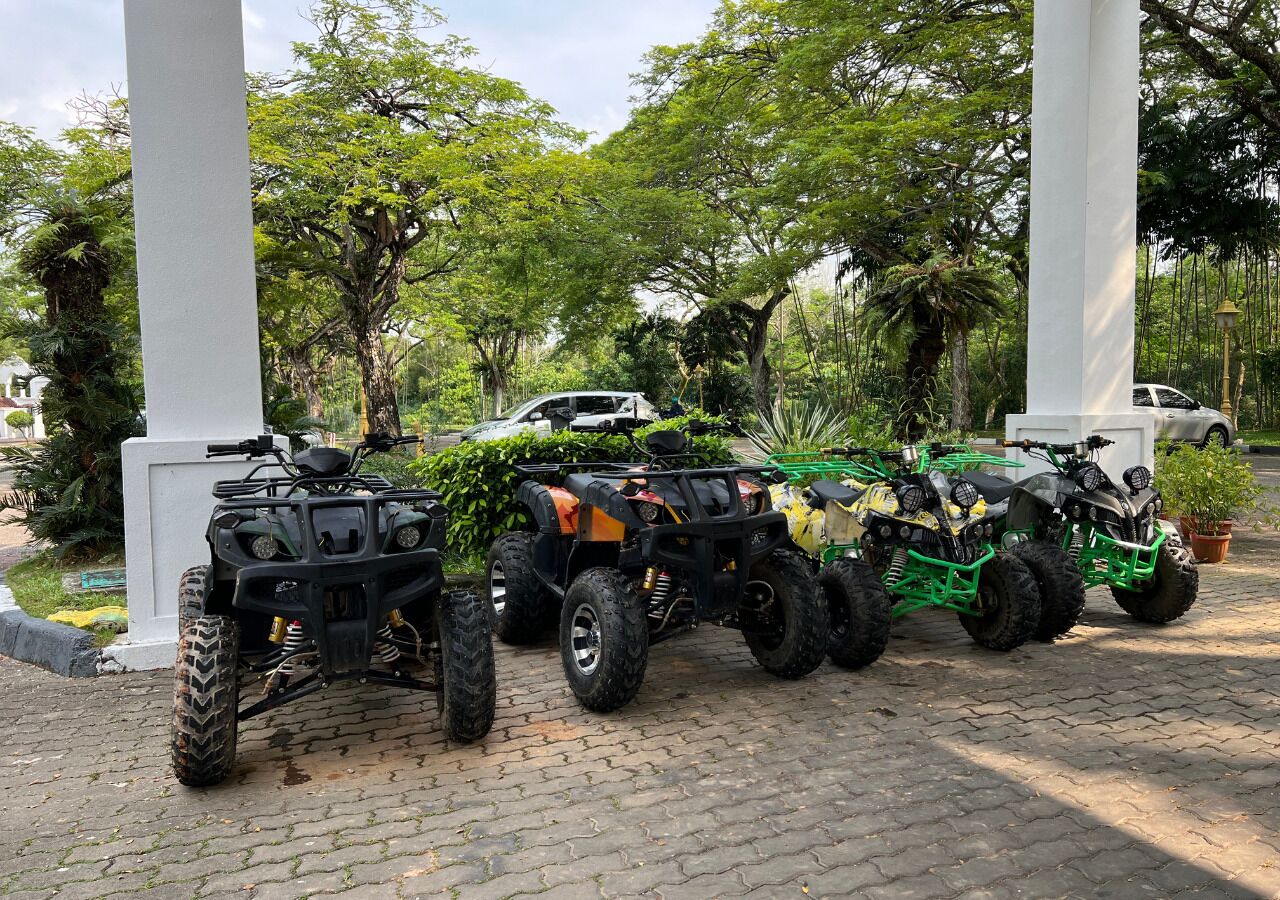 A row of ATVs parked on a paved area with trees and a white structure in the background.