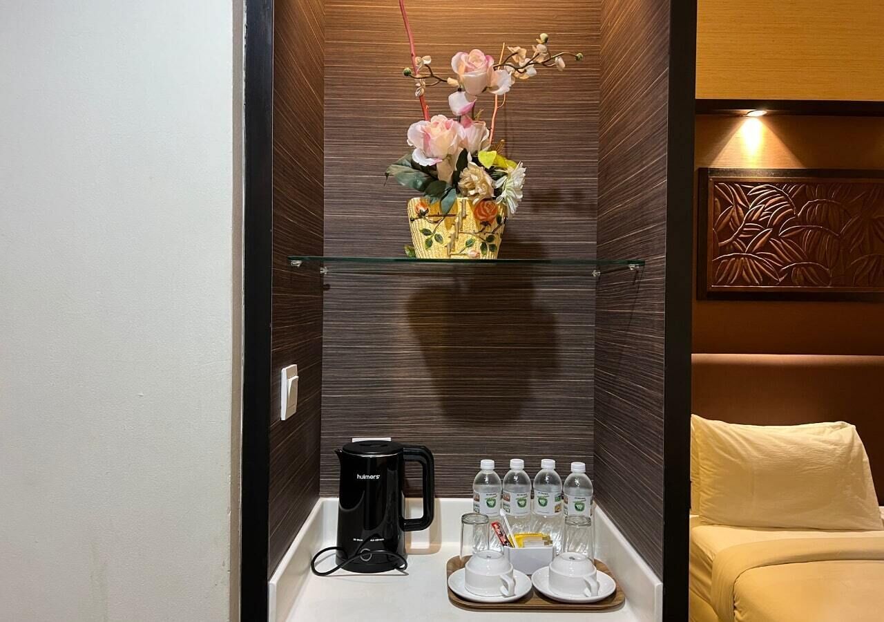 Quarto de hotel com minibar, cafeteira, vaso de vidro com flores e cama com travesseiro.