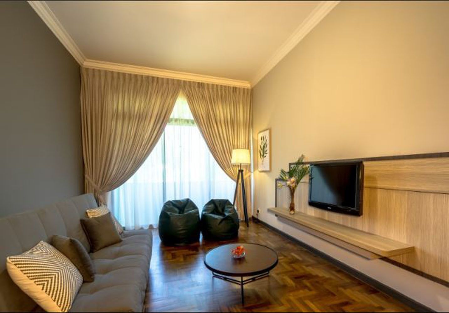 Une chambre d’hôtel avec un canapé, deux fauteuils poire, une table basse, un téléviseur fixé au mur et une grande fenêtre avec des rideaux.