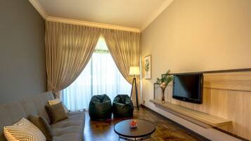 Une chambre d’hôtel avec un canapé, deux fauteuils poire, une table basse, un téléviseur fixé au mur et une grande fenêtre avec des rideaux.