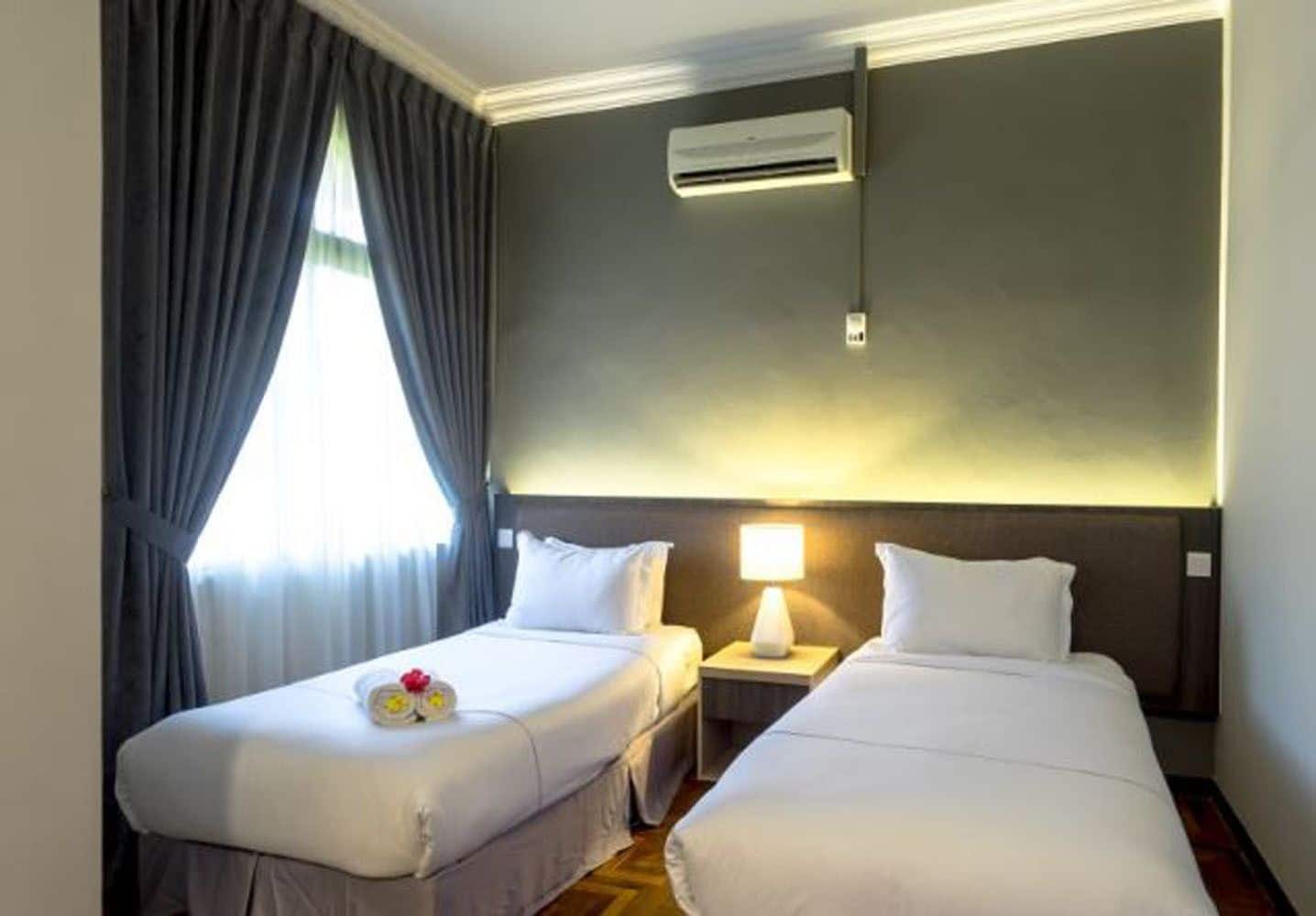 A hotel room with two beds, a nightstand with a lamp, an air conditioning unit, and a window with curtains.