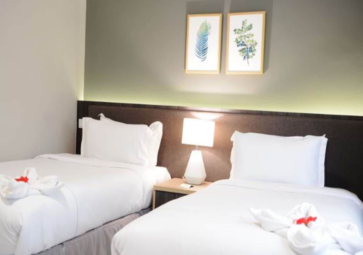 Habitación de hotel con dos camas, ropa blanca, una mesita de noche con lámpara e ilustraciones botánicas enmarcadas en la pared.