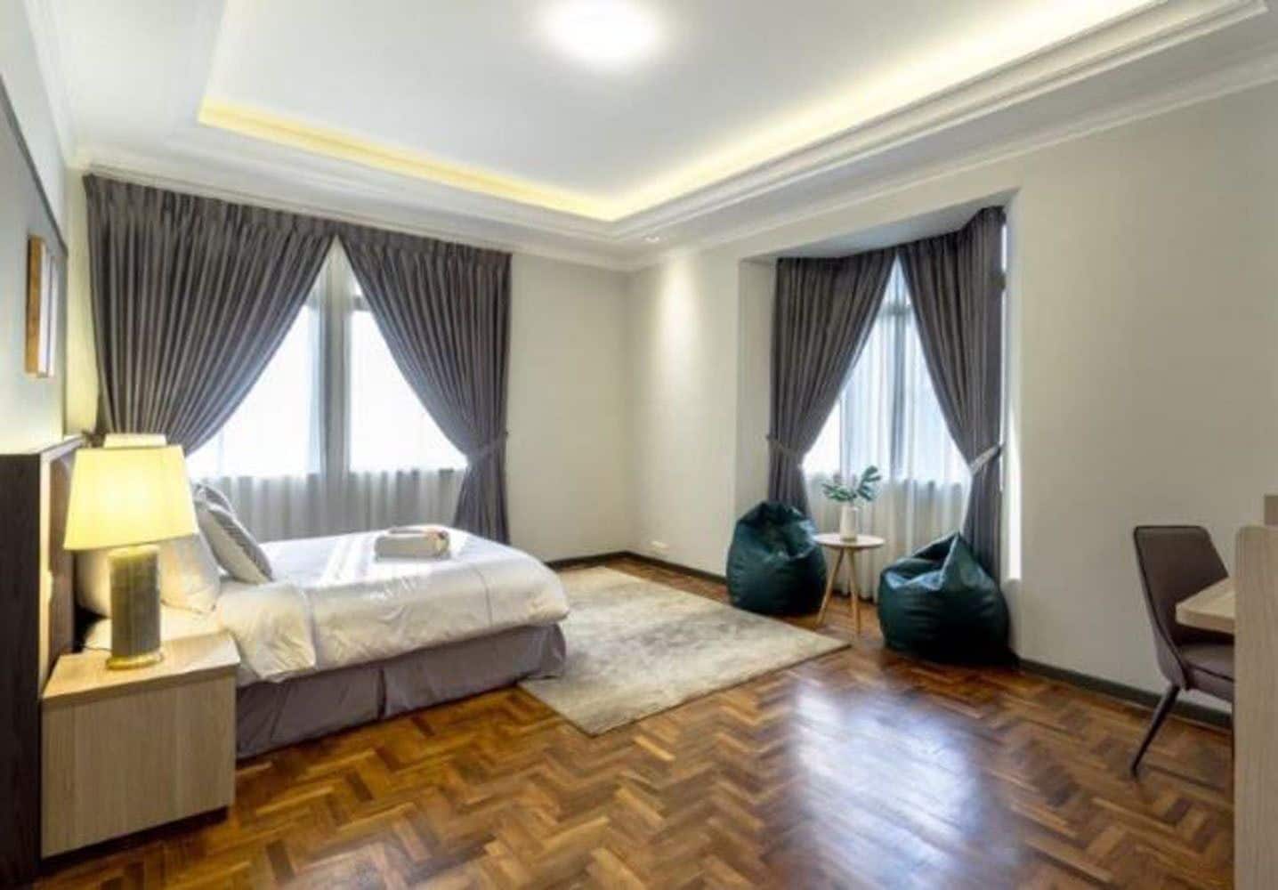 A modern hotel room with a wooden herringbone floor, a bed with a white duvet, a desk with a lamp, a green bean bag chair, and two windows with grey curtains.