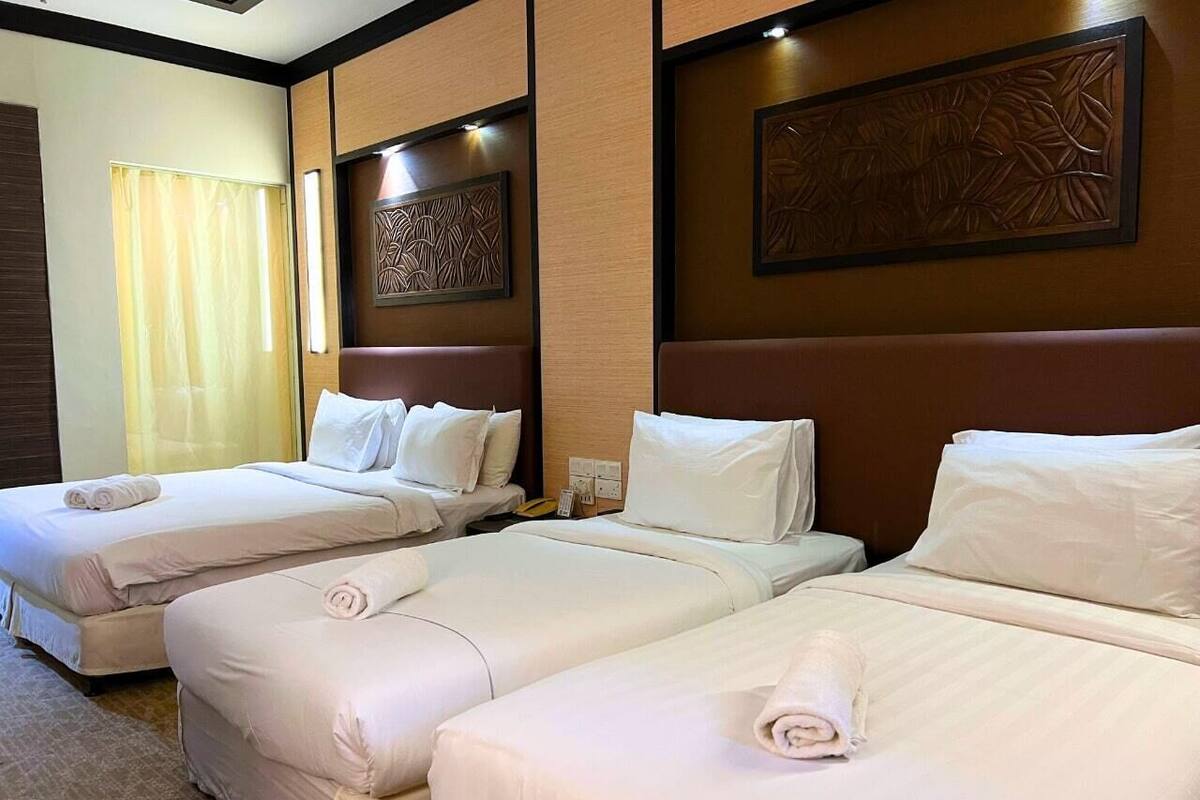 Habitación de hotel con tres camas, cada una con ropa de cama y almohadas blancas, cabeceros de madera con diseño floral y un ventanal con cortina.