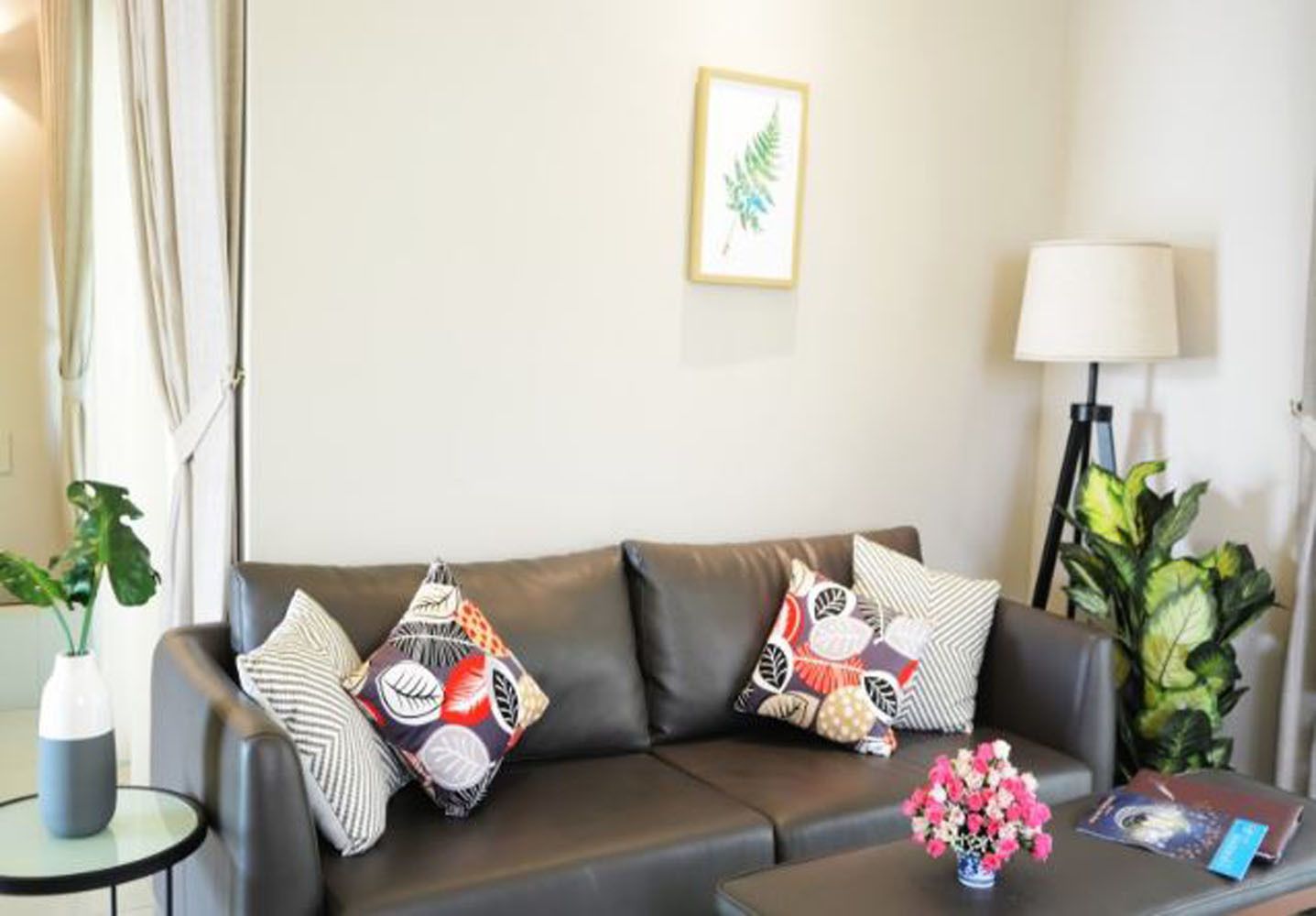 A modern living room with a leather sofa, patterned cushions, a framed plant, a floor lamp, and a small table with a vase of flowers.