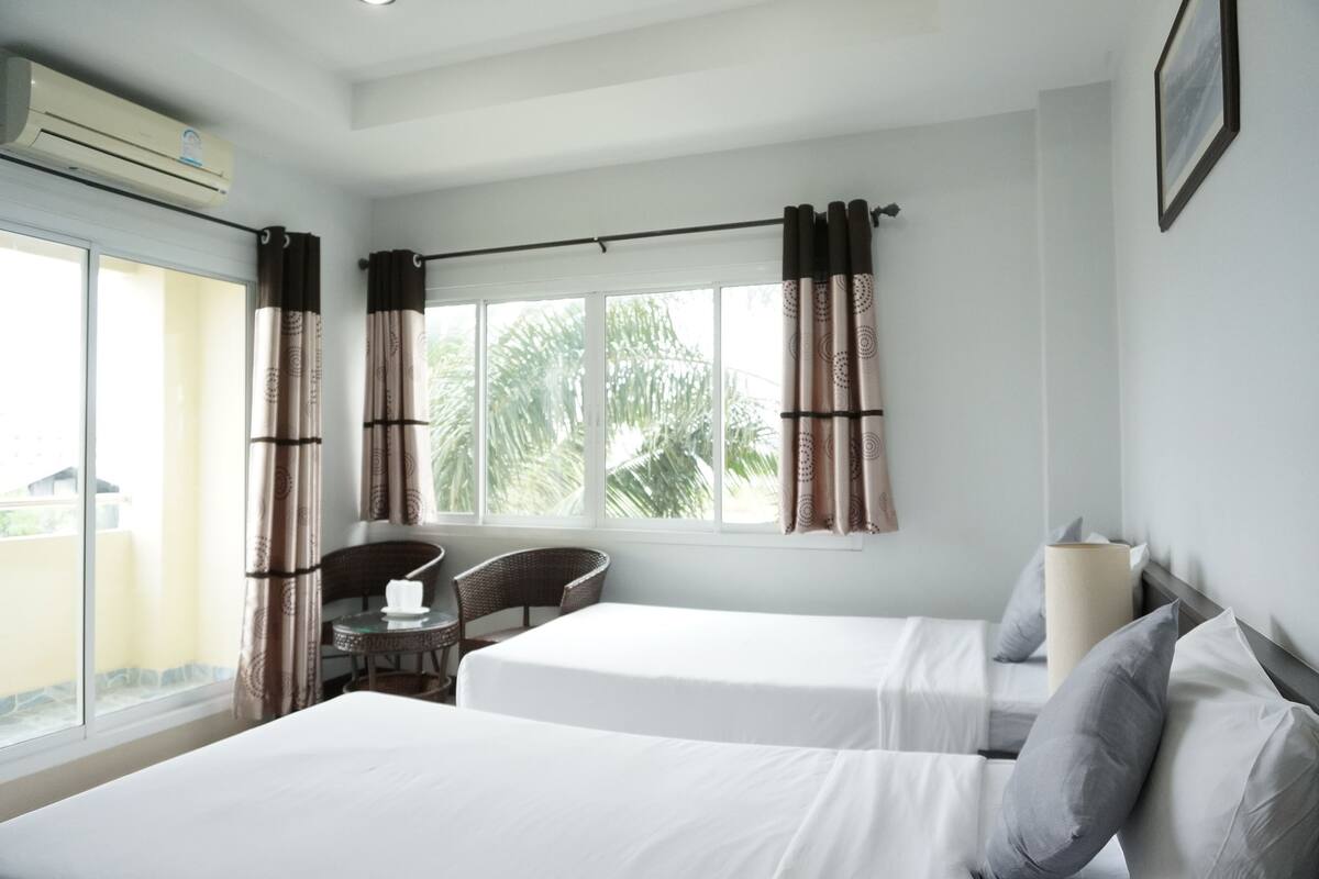 Ein Hotelzimmer mit zwei Betten, einem Fenster mit Vorhängen, einem Sessel, einem Tisch und einer Klimaanlage.