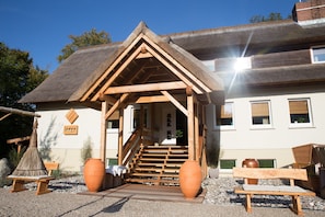 Une structure en bois surmontée d’un toit de chaume, avec un escalier en bois menant à une porte et un banc à l’extérieur.