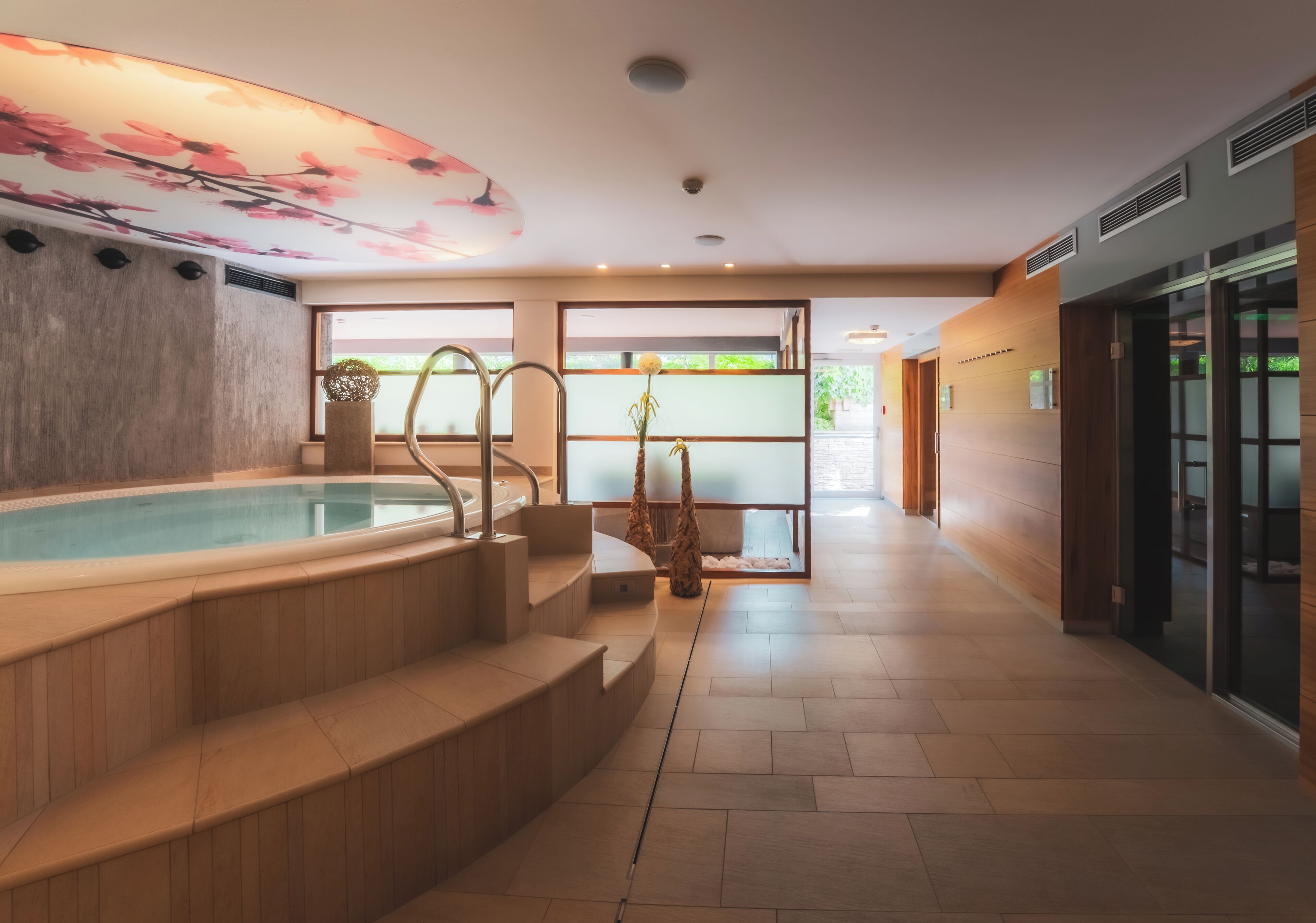 A modern indoor pool area with a wooden staircase leading to the pool, a glass door entrance, and a ceiling with a floral design.