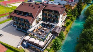 A multi-story building with a rooftop terrace, a river adjacent to the property, and a sports field in the background. - Hotel Kaprunerhof (Kaprun)
