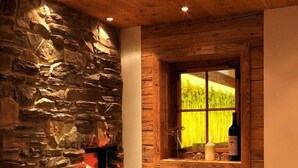 A cozy room with a stone wall, a wooden ceiling, a fireplace, a window with a view of greenery, and a wooden stool with a bowl of fruit. - Hotel Kaprunerhof (Kaprun)