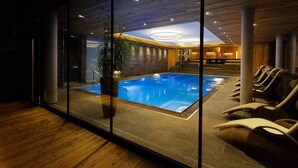 A modern indoor swimming pool with a glass wall, wooden ceiling, and lounge chairs. - Hotel Kaprunerhof (Kaprun)