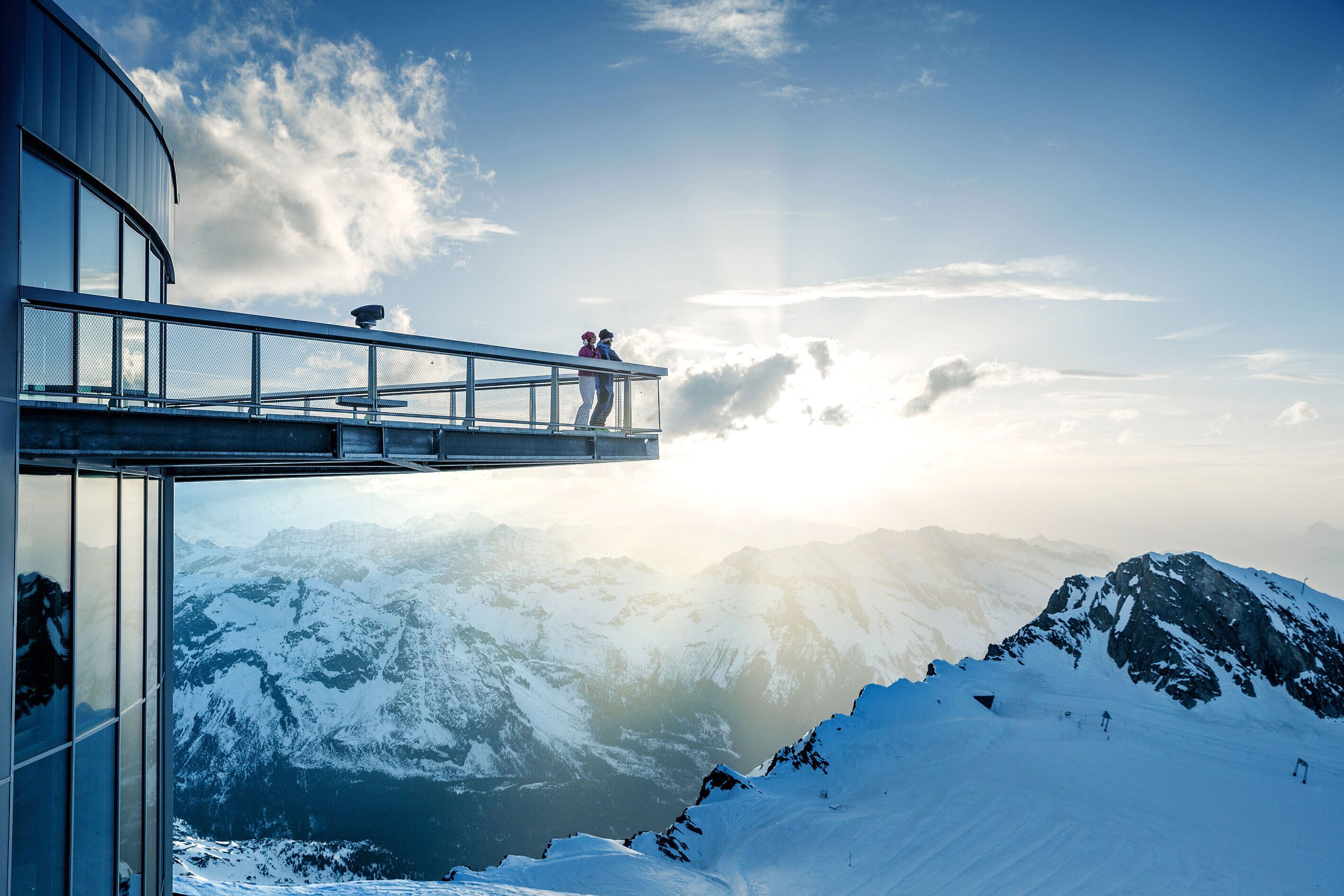 Un balcón de cristal con vistas a un paisaje montañoso nevado y cielo despejado.