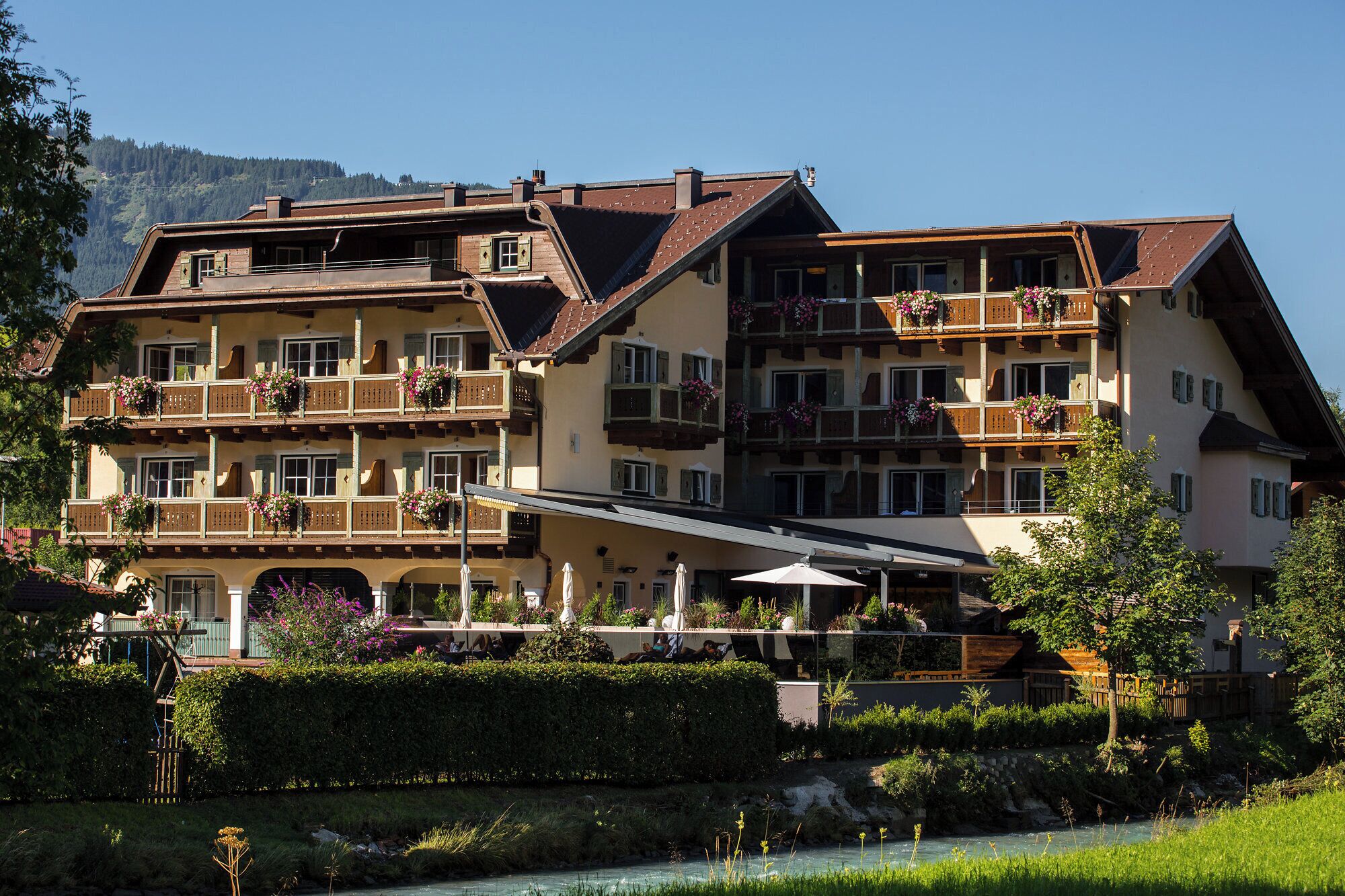 Een hotel met meerdere verdiepingen, balkons, een rivier ervoor en een bergachtig achtergrondlandschap.