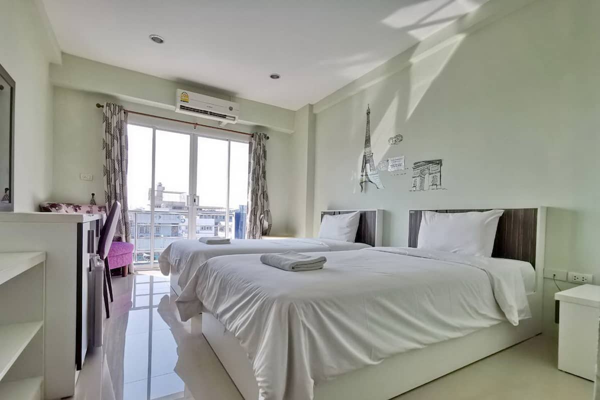 A modern hotel room with two beds, a large window with curtains, an air conditioning unit, and a view of the cityscape.