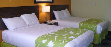 A hotel room with two beds, each with white bedding and green floral bedspreads. There is a framed picture on the wall above the beds, a bedside lamp, and a telephone on the nightstand.