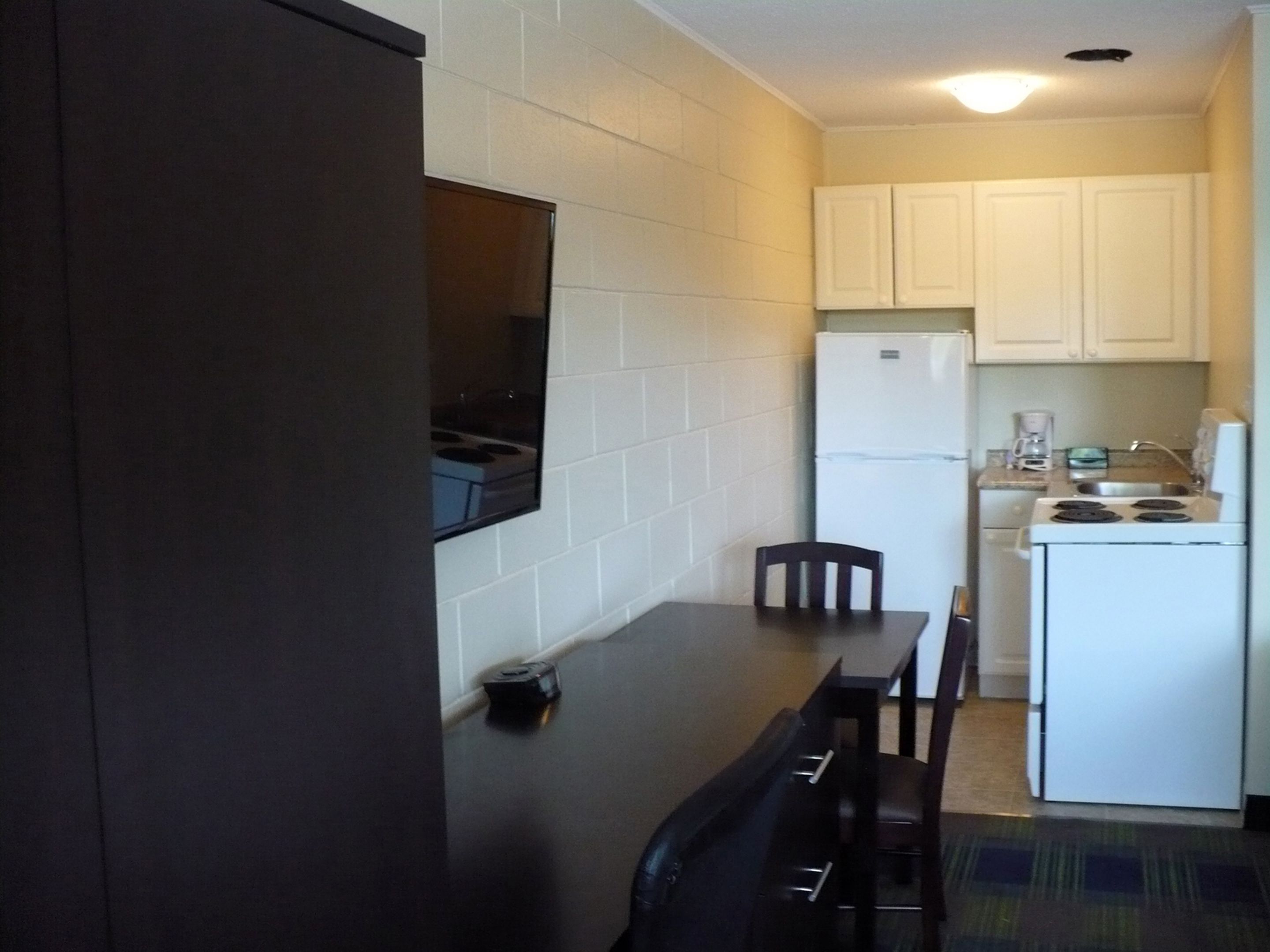 A compact kitchen with white cabinets, a white refrigerator, a stove, and a microwave. There is a small table with a chair, and a dark-colored bench.