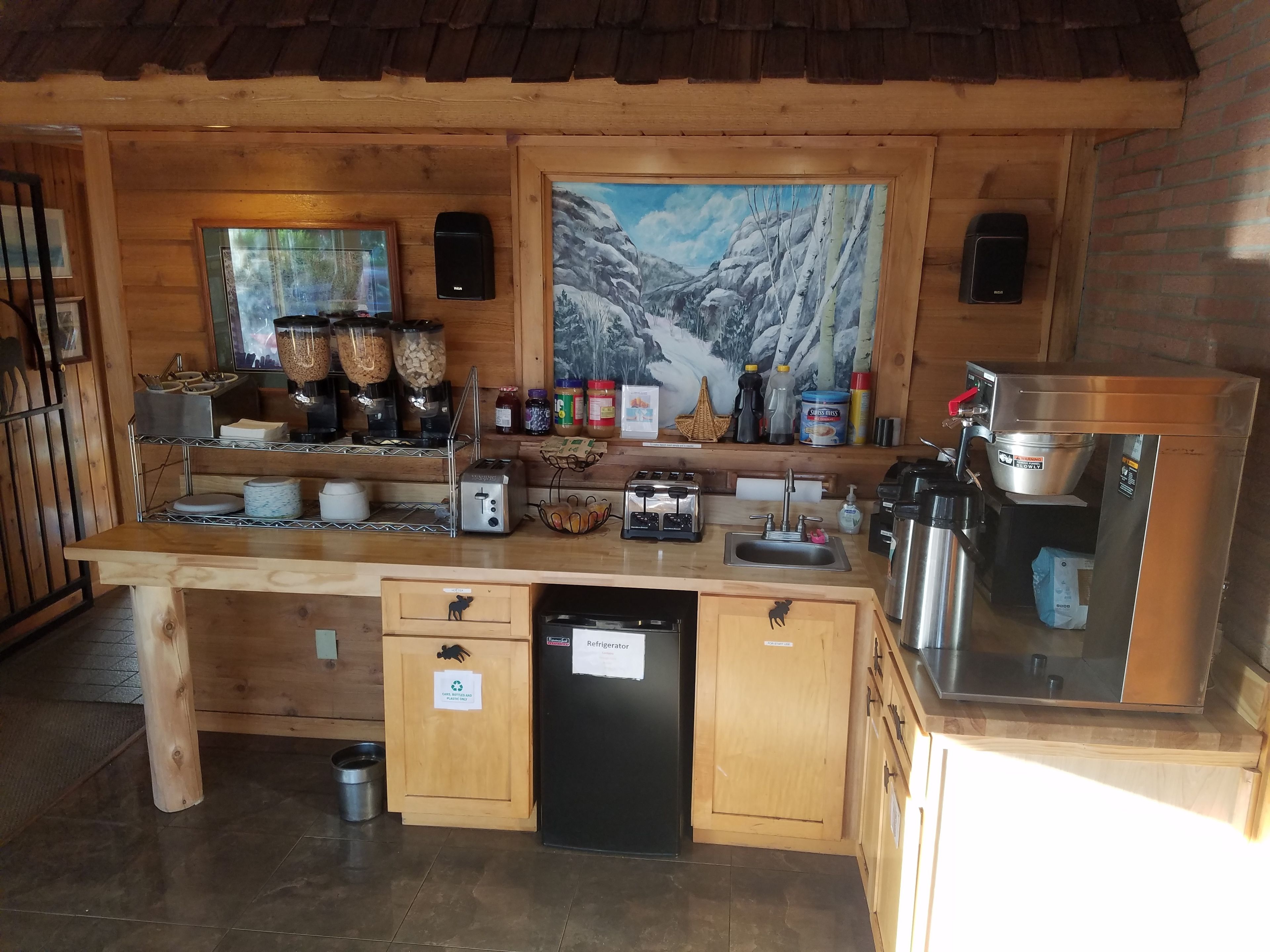 A kitchen with wooden cabinets, a refrigerator, a coffee machine, and a painting of a snowy mountain.