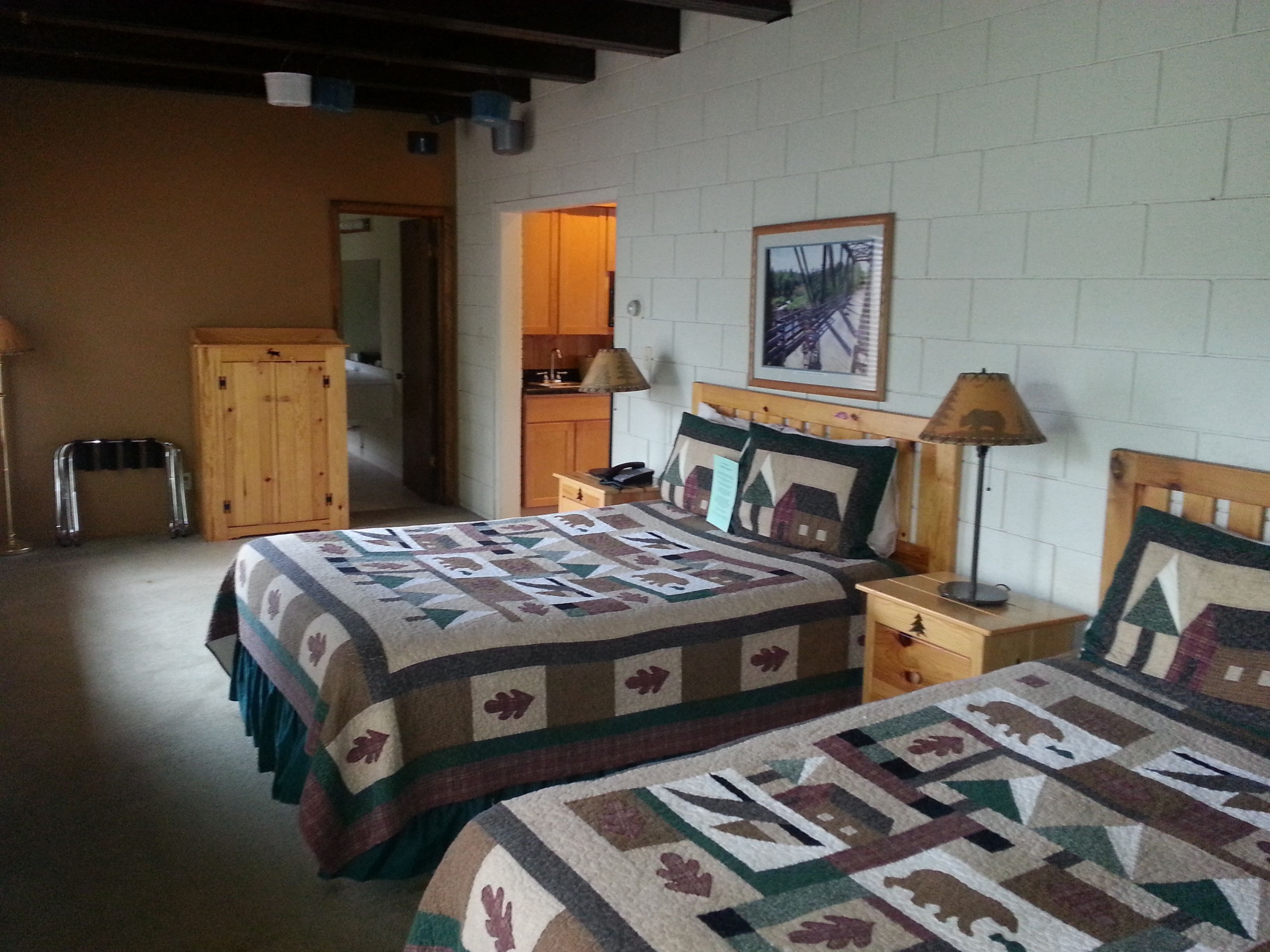 Un dormitorio con dos camas, cabecero de madera, mesita de noche, lámpara, un cuadro enmarcado en la pared y una cocina visible a través de una puerta abierta.