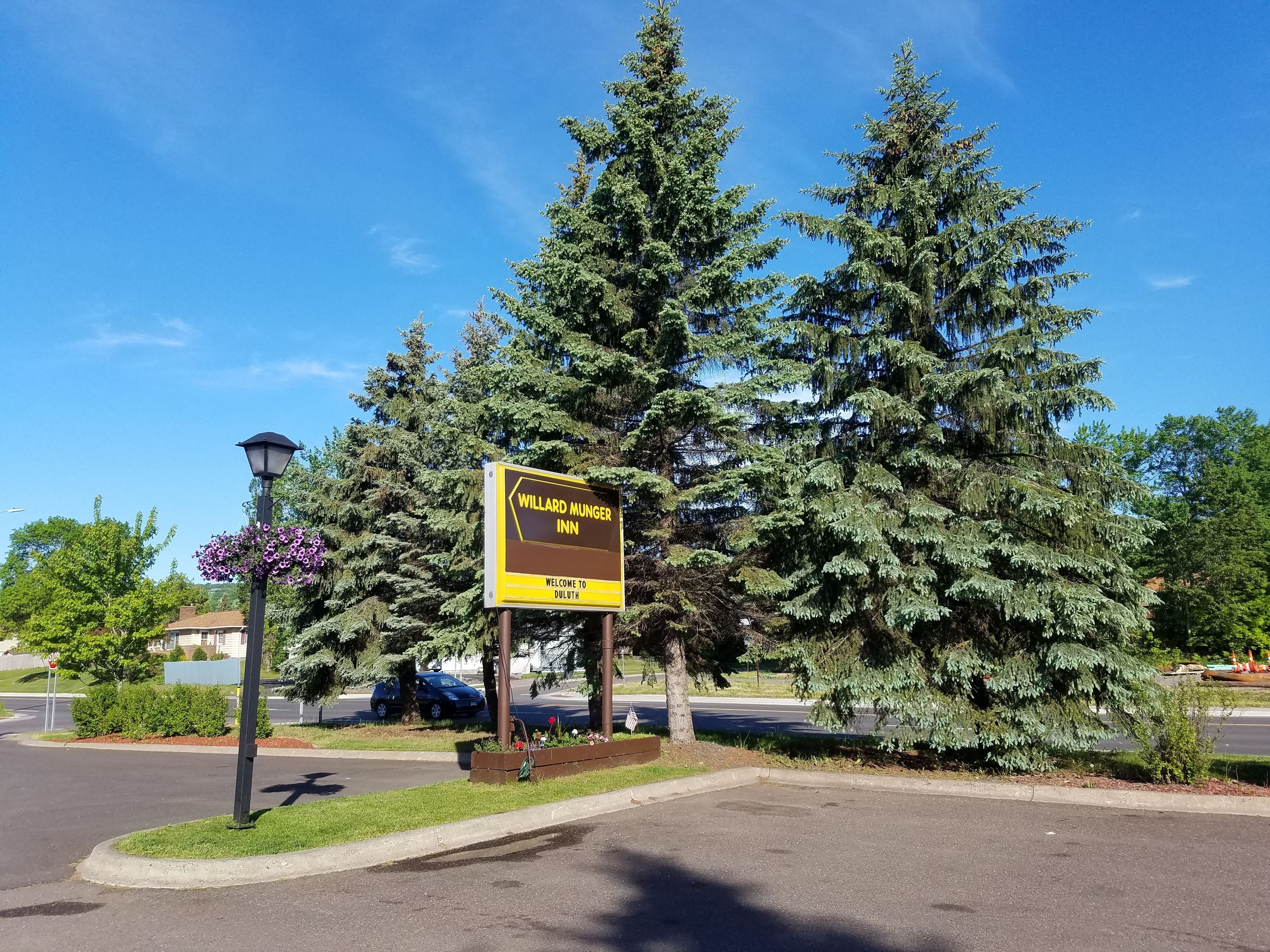 Une enseigne pour le Willard Hunger Inn, avec un message d’accueil à l’intention de Duluth, entourée d’une végétation luxuriante et sous un ciel bleu clair.
