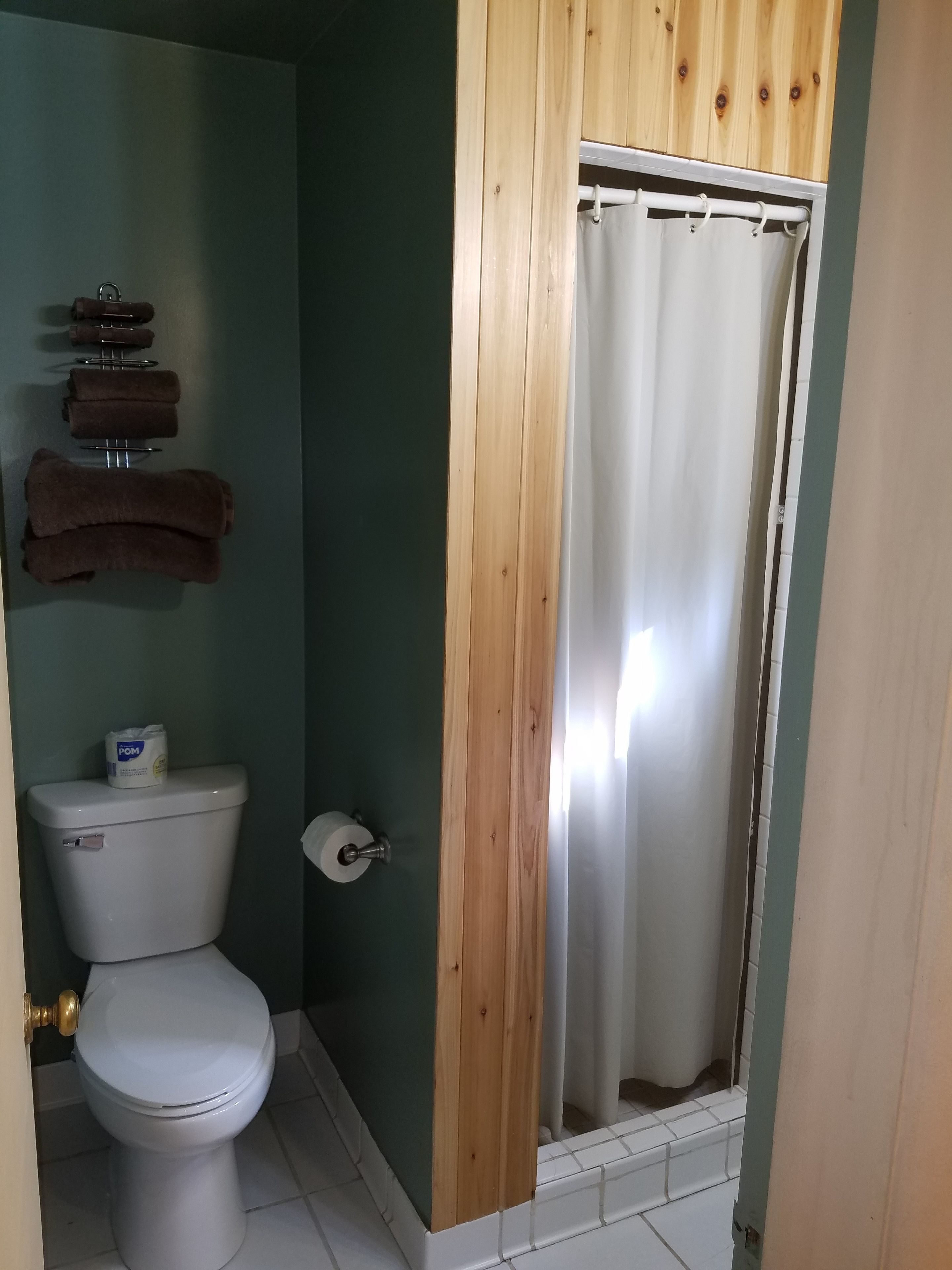 A bathroom with a toilet, a wooden panel, a shower curtain, and a towel rack with towels.