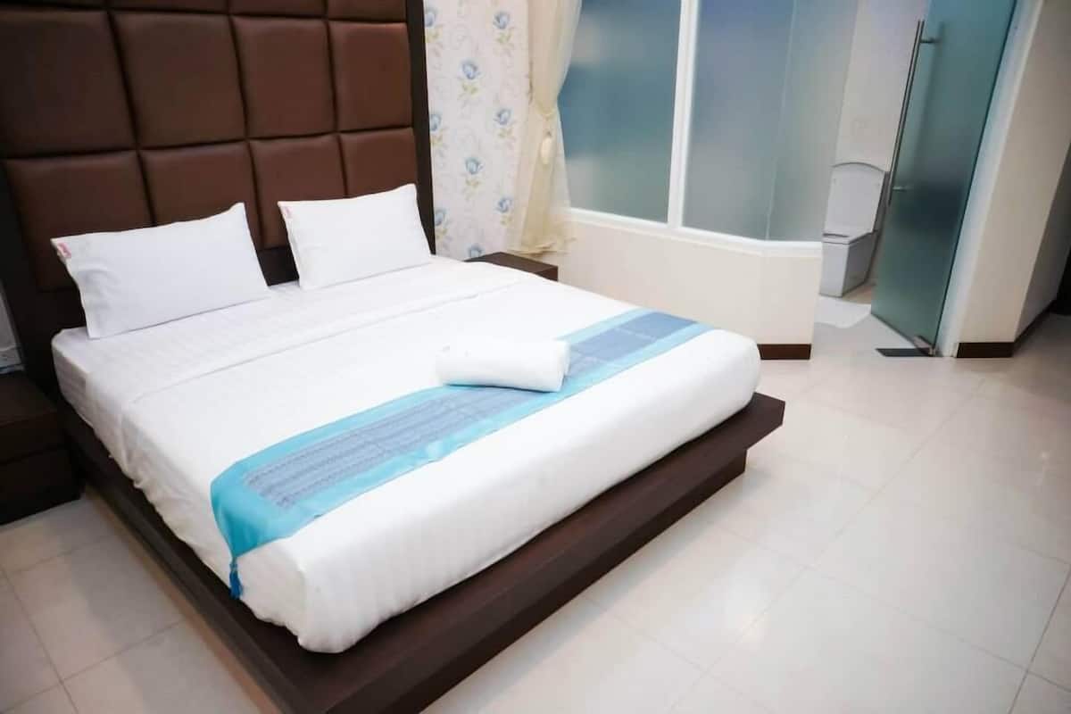 A neatly made bed with white linens and a blue accent, a brown headboard, and a window with frosted glass.