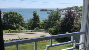 En balkon med to hvide metalstole og et lille rundt bord, med udsigt over vand med både og en fjerntliggende ø.