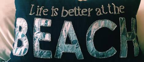 Ein Kissen mit der Aufschrift "Life is better at the BEACH" auf einem Bett mit einer blauen, gesteppten Bettdecke.