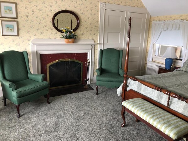 A bedroom with a four-poster bed, two green armchairs, a fireplace, and a mirror on the wall.