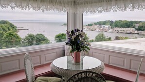 Et værelse med et rundt glastæppe, to fletstole, en blomstervase og udsigt over en havn med både og bygninger.