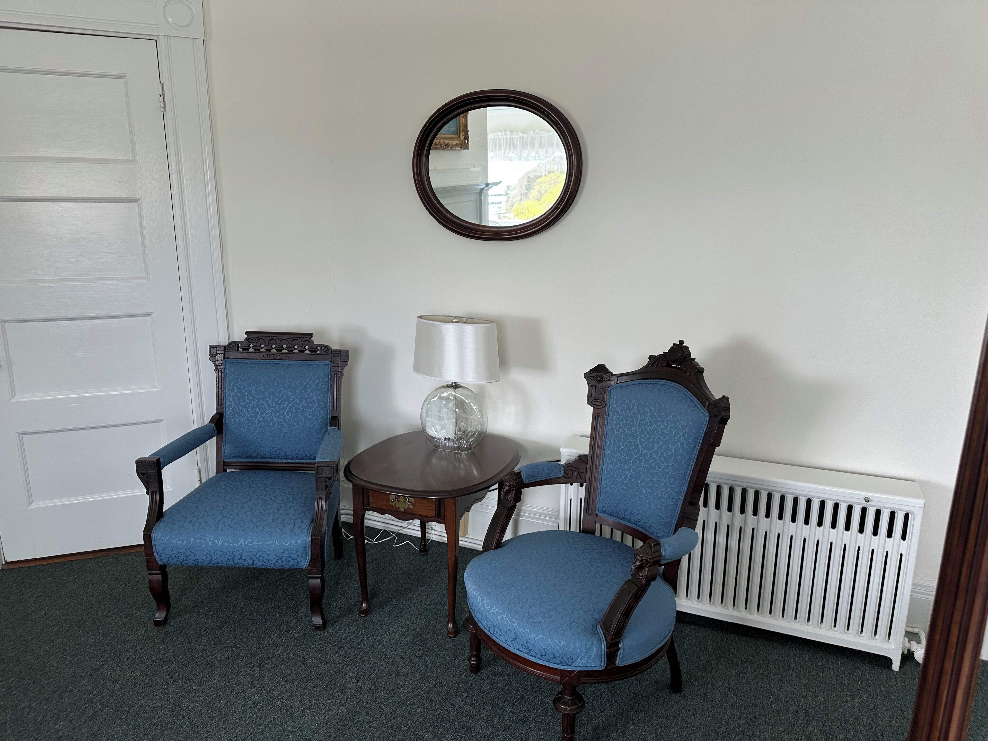 A room with two blue upholstered chairs, a small round table with a lamp, and a round mirror on the wall.