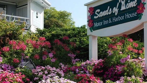 Et skilt for "Sands of Time Motor Inn & Harbor House" omgivet af blomstrende lyserøde og lilla blomster.