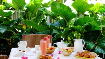 Une table dressée pour le petit-déjeuner, avec une carafe, des tasses, un saladier de fruits et des viennoiseries, le tout entouré d’un luxuriant feuillage verdoyant.