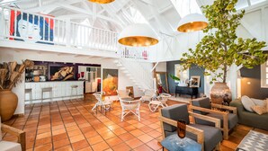 A spacious lobby with a high ceiling, white railings, and a staircase. There are several seating areas with chairs and tables, a large vase with plants, and artwork on the walls. - Hôtel Plein Soleil (Le Francois)