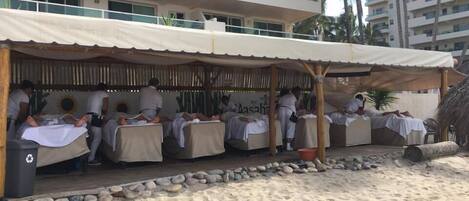 A beachside massage area with multiple massage tables under a canopy, a stone border, and a palm tree in the background.