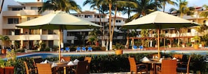 Un resort con terraza al aire libre cerca de una piscina, rodeado de palmeras y un edificio con varios balcones.