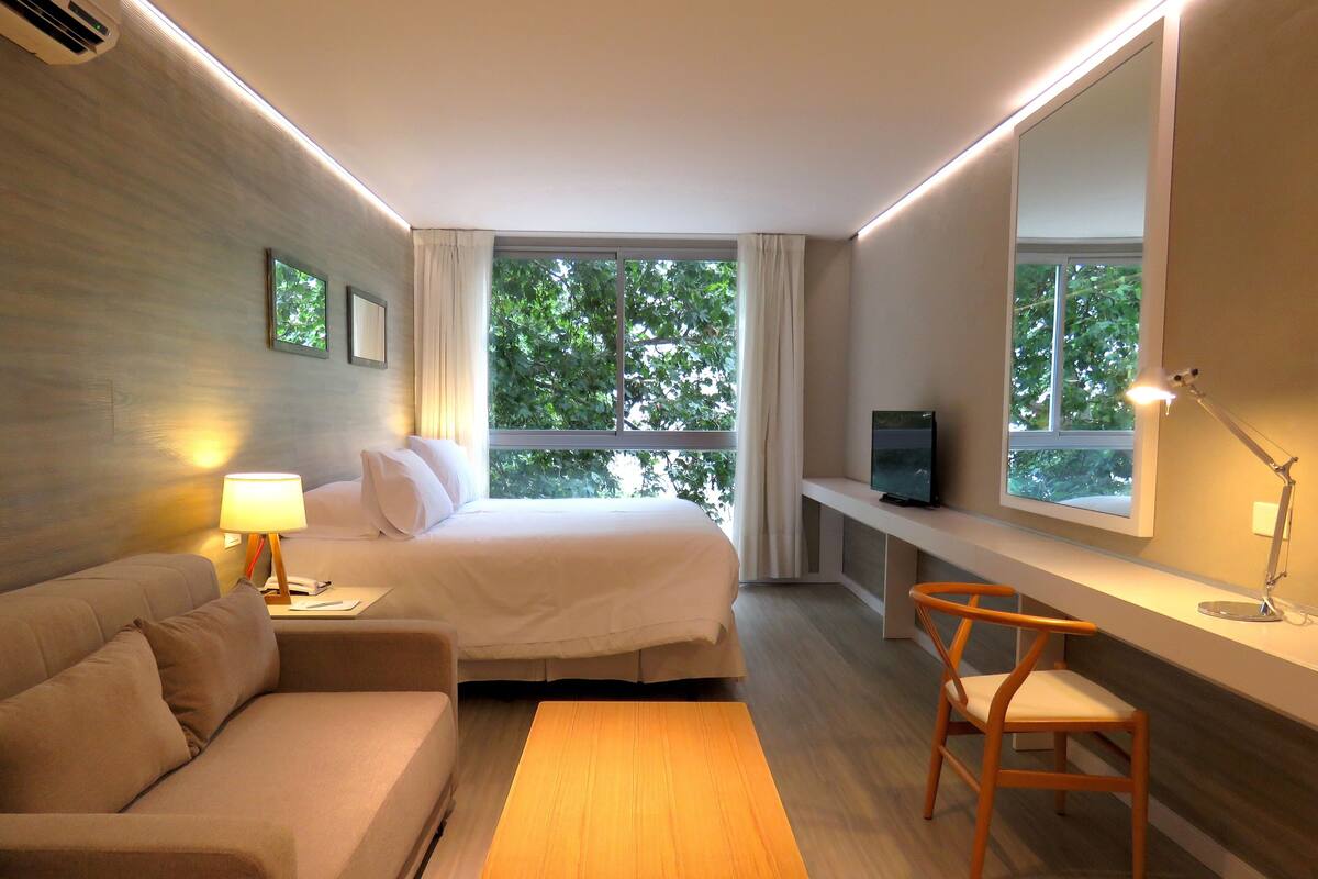 Quarto de hotel com uma cama, um sofá, uma escrivaninha, uma televisão e uma janela com vista para árvores.