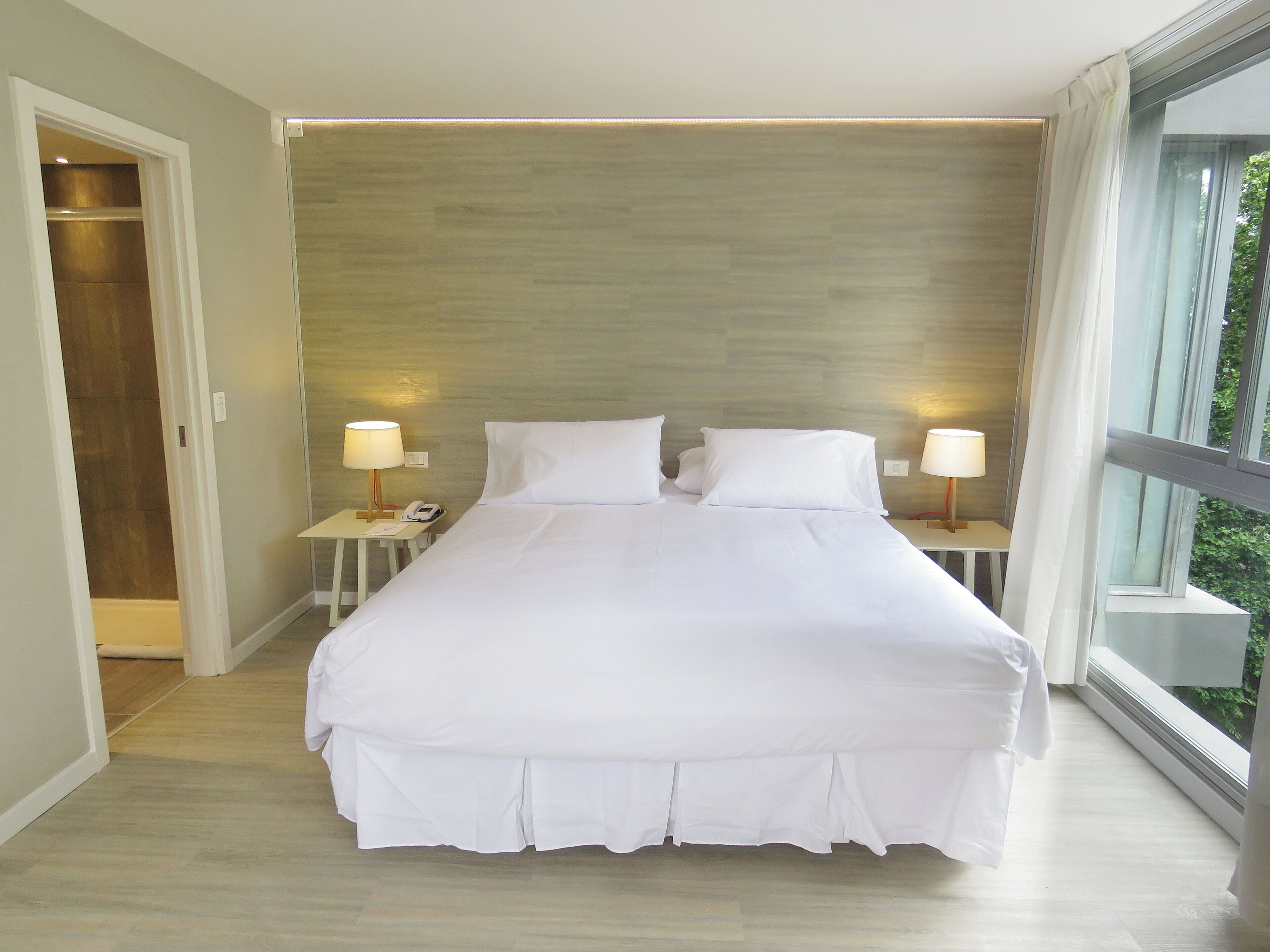 Habitación de hotel con una cama grande, dos mesitas de noche con lámparas, un cabecero de madera y vistas a través de un ventanal amplio.